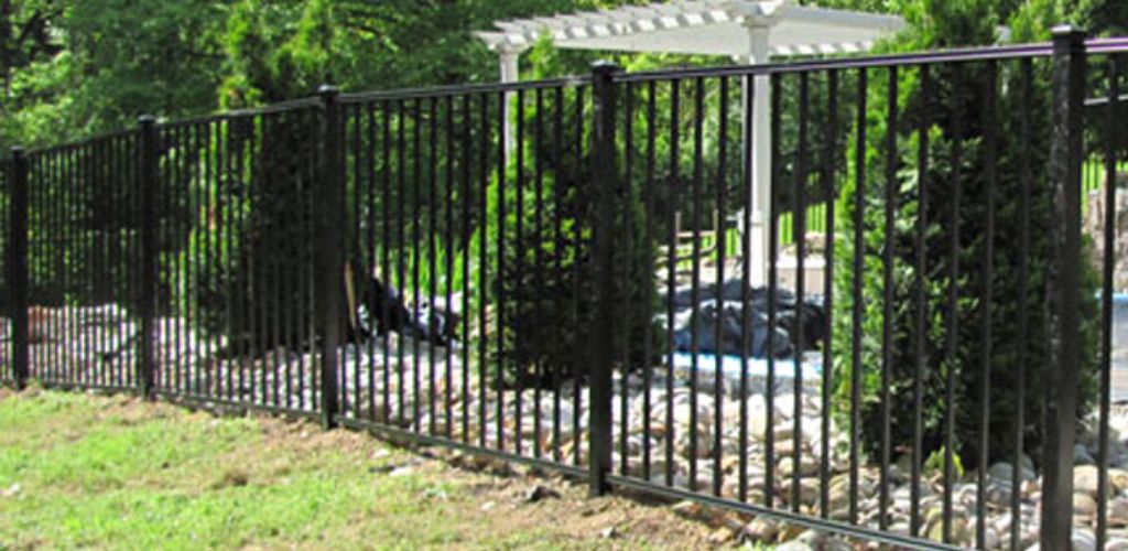 A black metal fence style surrounds a swimming pool in a backyard with trees and shrubs.